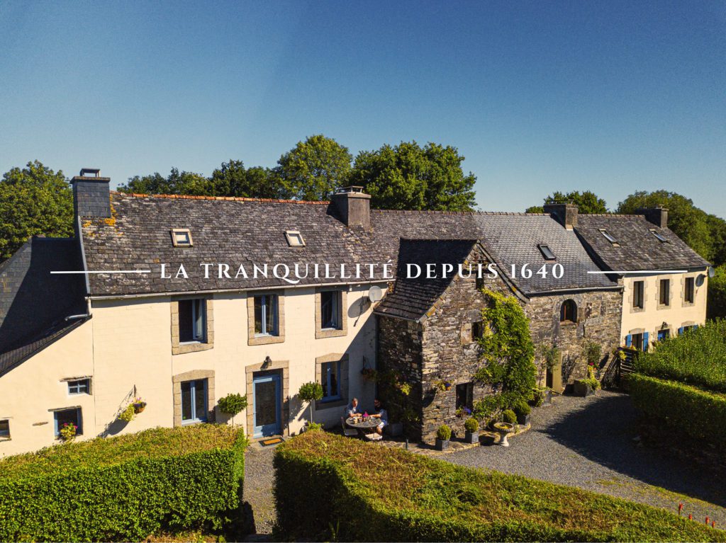 Kergudon Gîtes séjour en Bretagne montrant la façade des bâtiments principaux avec un ciel bleu et un couple d'homosexuels assis sur la terrasse du petit déjeuner.